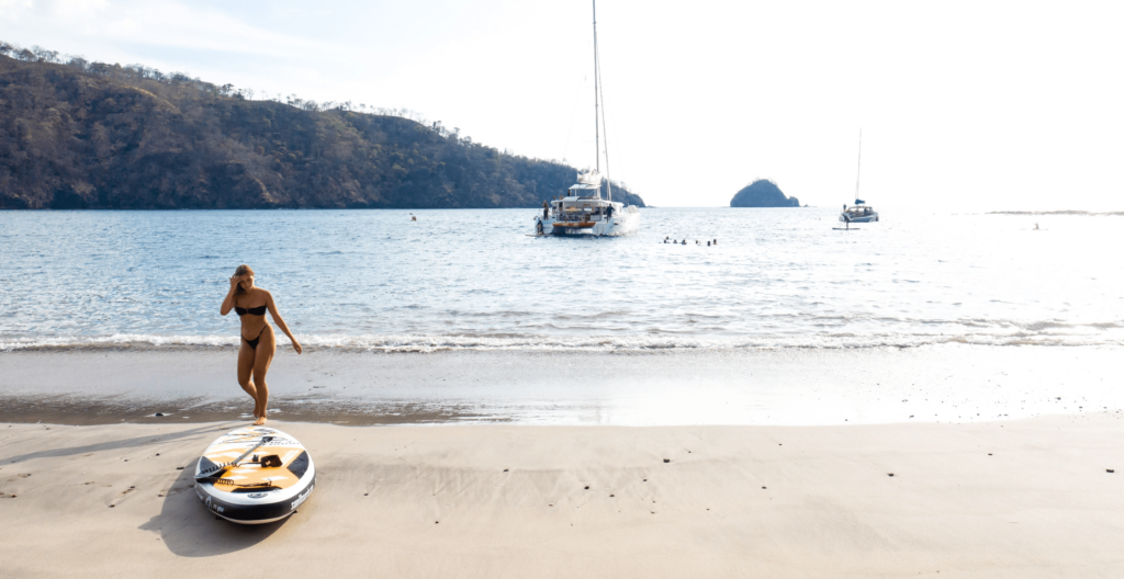 A woman in a bikini walks along the beach beside a paddleboard, with sailboats anchored in the background and hills in the distance