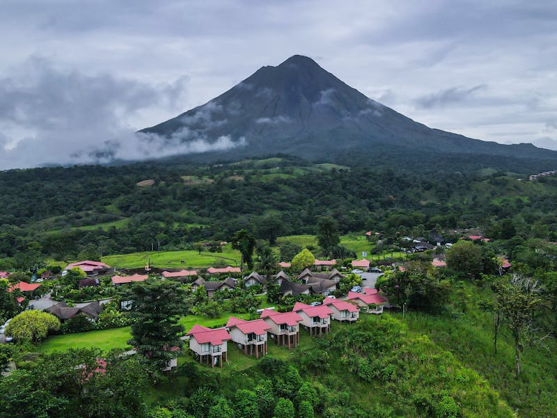  Arenal Volcano rises majestically in the background, partially covered by clouds, with a cluster of lodges in the foreground surrounded by lush greenery.

