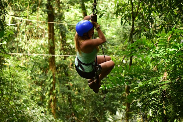 A person ziplining through a lush green jungle, wearing a helmet and harness, surrounded by dense foliage.

