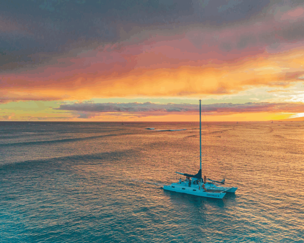 A sailboat on calm waters during sunset, with vibrant orange and purple skies reflected in the ocean.